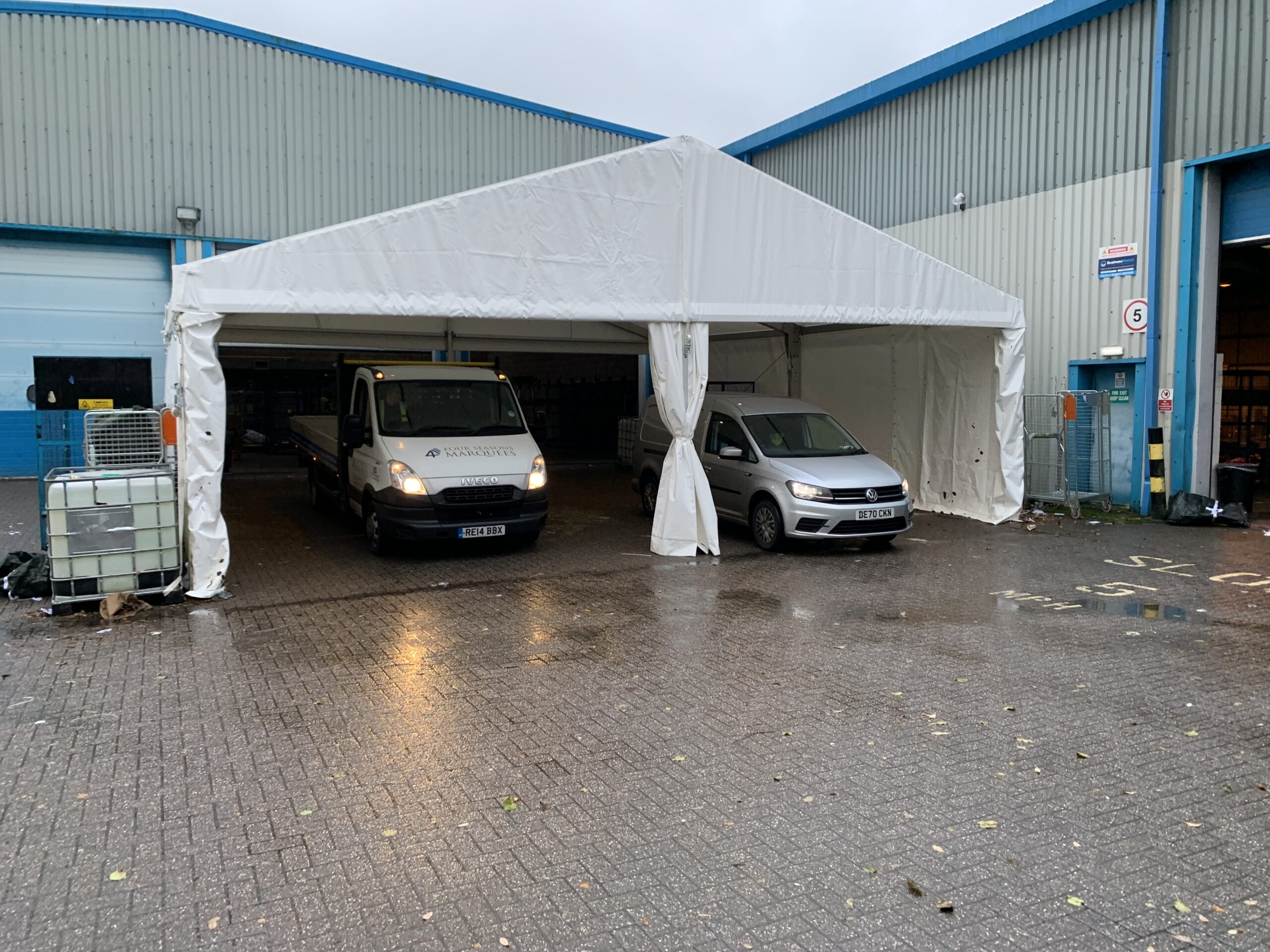 Temporary storage marquee providing covered parking and weather protection for commercial vans and cars beside an industrial warehouse.