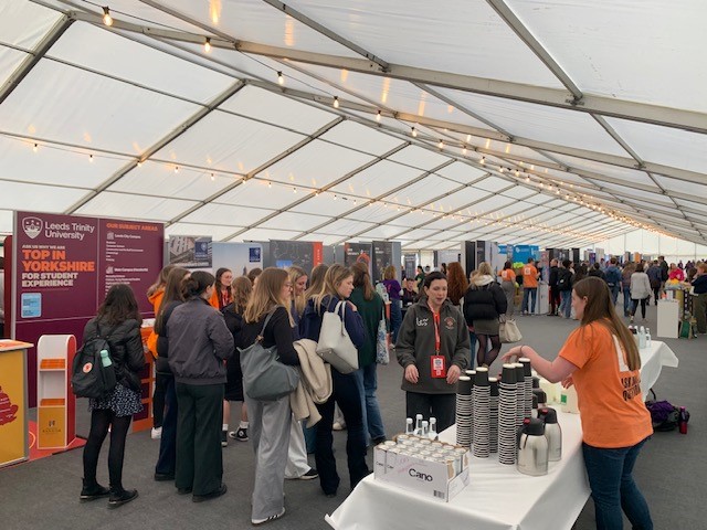 Busy indoor exhibition inside a clearspan marquee with visitors gathering around display stands and product tables.